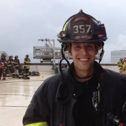 Tyler Capron smiling in a Boulder Fire Rescue helmet and turnout coat, with firefighters on a rooftop behind