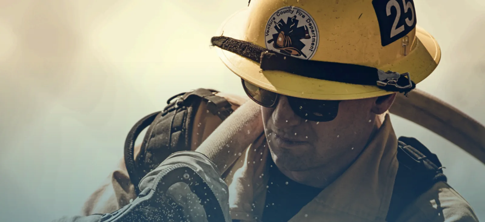 Firefighter in a yellow helmet and sunglasses carries a fire hose, with water droplets in the air