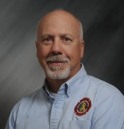 Jeff Dill, a gray-bearded man in a light blue uniform shirt with a fire department patch, against a gray background