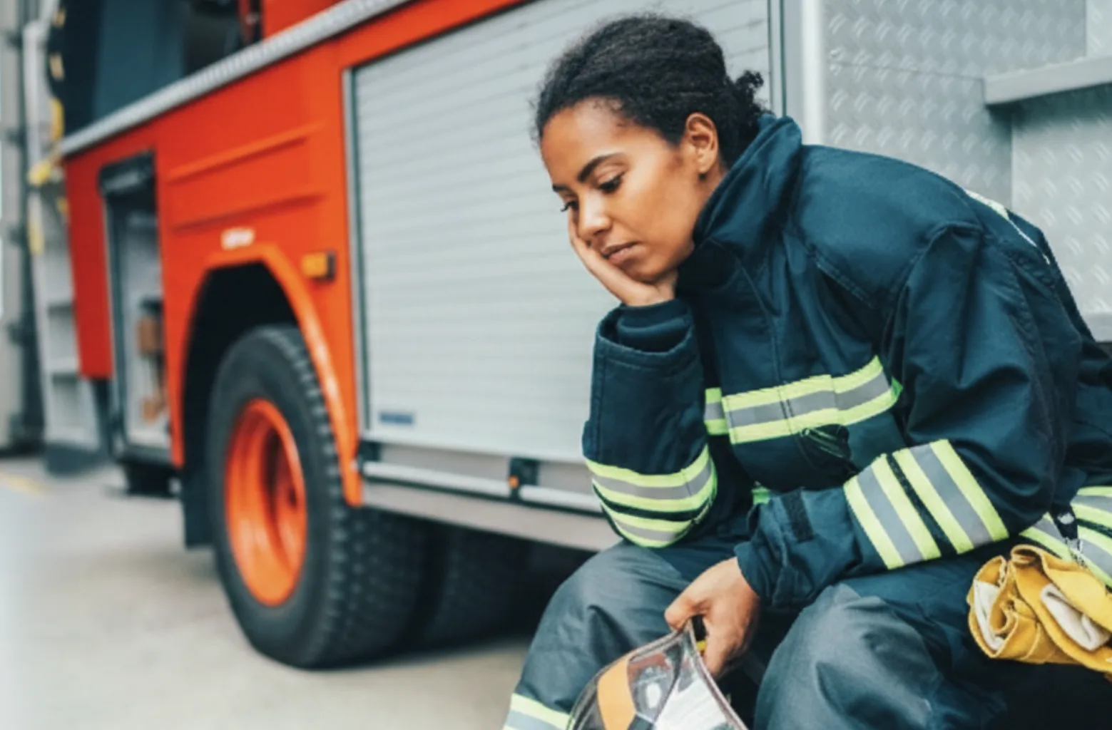 Woman firefighter in turnout gear sits by a red fire engine, resting her chin on her hand and looking down