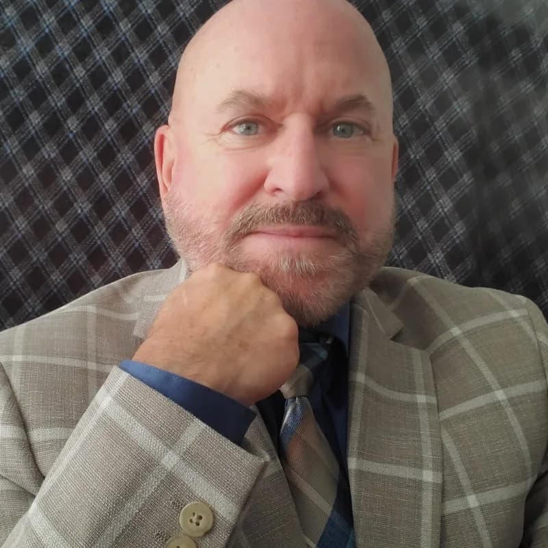 Robert Avsec, a bald man with a beard in a plaid jacket and tie, resting his chin on his fist