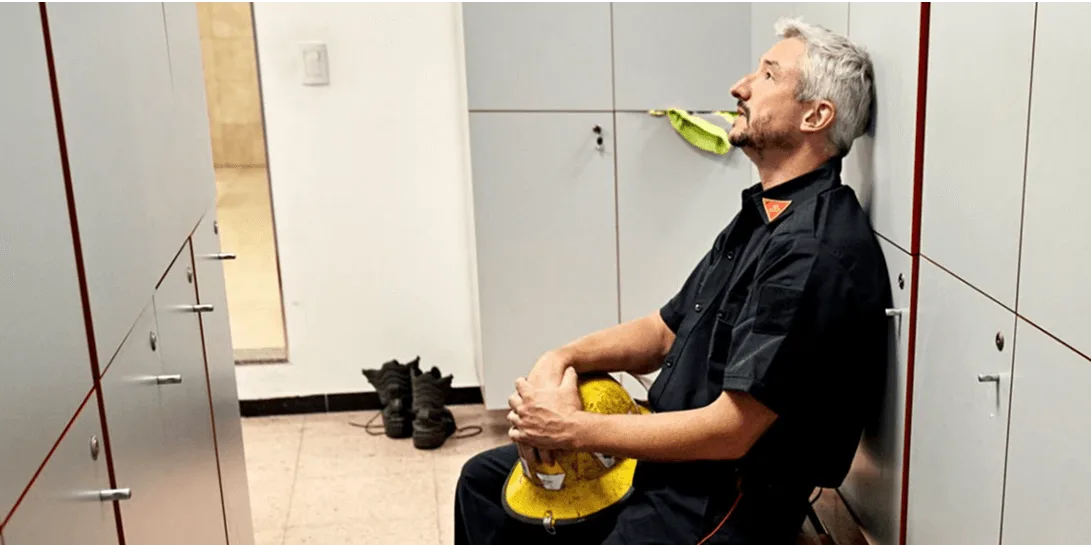 Firefighter sits in a locker room with a yellow helmet in his lap, looking up at the ceiling
